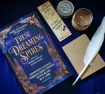 photograph showing a copy of These Dreaming Spires, edited by Marie O'Regan and Paul Kane,lying on a purple surface. To the right of the book are a These Dreaming Spires bookmark, an In These Hallowed Halls jar candle with the lid off, a beige notebook and a white feather quill pen
