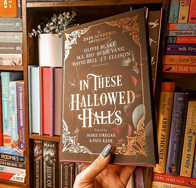 a woman's hand holds a copy of In These Hallowed Halls, edited by Marie O'Regan and Paul Kane, up against rows of bookshelves