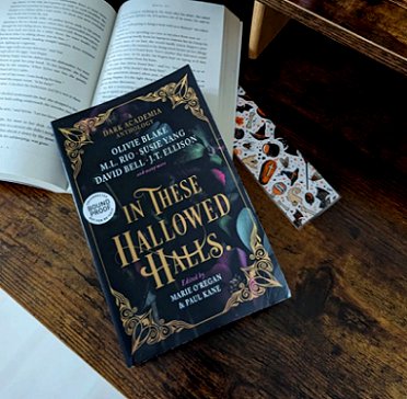 Image showing a copy of In These Hallowed Halls, edited by Marie O'Regan and Paul Kane, lying on top of an open book alongside a bookmark, on a wooden surface