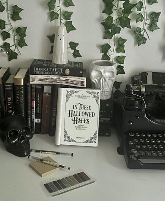 image showing a copy of In These Hallowed Halls, edited by Marie O'Regan and Paul Kane, standing open to the title page, leaning against a row of books. In the foreground is a paint strip, two pens and a post-it pad in front of a black skull. TO the right is a black typewriter, and standing on top of the books at the back are a ghost statue and a skull ornament. Ivy leaves are dropping down the wall at the back