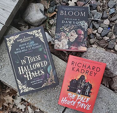 photograph showing three books lying on a plank and pebbles. The books are In These Hallowed Halls, edited by Marie O'Regan and Paul Kane, The Pale House Devil by Richard Kadrey, and Boom by Delilah S. Dawson