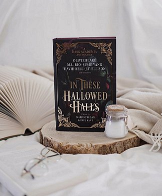 photograph of a copy of In These Hallowed Halls, edited by Marie O'Regan and Paul Kane, standing on a piece of a log, alongside an open book with pages fanning out, a small corktopped glass jar of milk and a pair of glasses, laying on a beige blanket on a white cloth and a white background