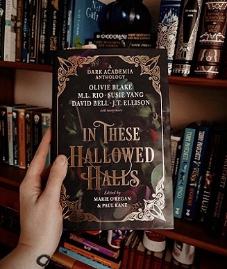 photograph showing a hand holding a copy of In These Hallowed Halls, edited by Marie O'Regan and Paul Kane, up against a full bookcase