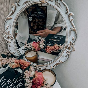 Image of an ornate white mirror on a whitle wall, reflecting the image of a woman dressed in a white shirt, holidng up a copy of In These Hallowed Halls, edited by Marie O'Regan and Paul Kane. A candle, pink flowers, and a copy of If We Were Villains by ML Rio feature in the foreground