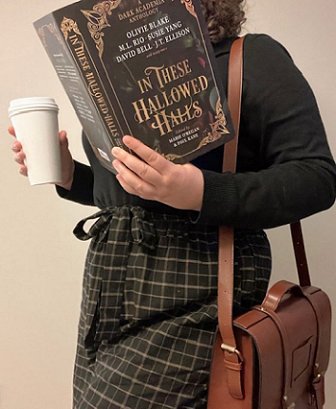 photograph of a woman in a black longsleeved top and black and white check trousers holding a styrofoam cup in one hand and a copy of In These Hallowed Halls, edited by Marie O'Regan and Paul Kane, in the other. A brown handbag hangs over one shoulder