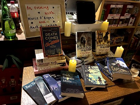 books displayed on a wooden table in front of a sign reading An evening with Marie O'Regan and Mark A. Latham. Titles featured include Death Comes at christmas, Dark Satanic Mills, The Mammoth Book of Ghost Stories by Women, Wonderland, Cursed, The Secret Romantic's Book of Magic, In Times of Want, Resurrection Blues, Bury Them deep, Thread of the Infinite, The Last Vigilant, and two Sherlock Holmes novels
