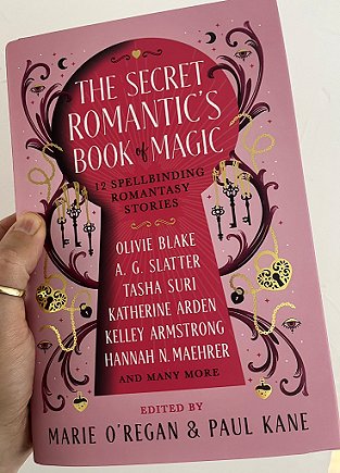 photograph of a man's hand holding a copy of The Secret Romantic's Book of Magic, edited by Marie O'Regan and Paul Kane, against a white background