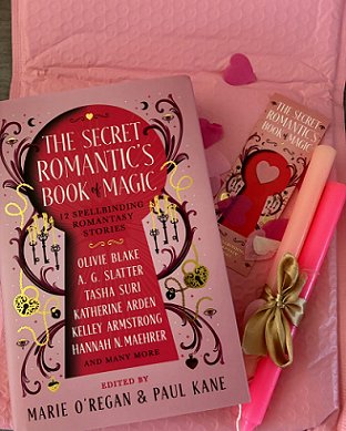 photograph of a copy of The Secret Romantic's Book of Magic, edited by Marie O'Regan and Paul Kane, on top of a pink padded envelope and some pink heartshaped confetti, alongside a Secret Romantic's bookmark and two pink candles tied with a gold ribbon