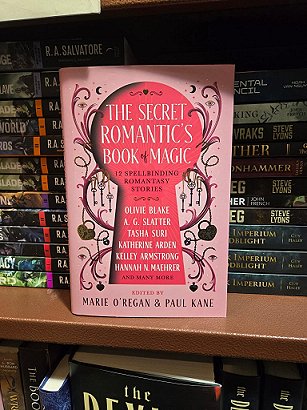 photograph showing a copy of The Secret Romantic's Book of Magic, edited by Marie O'Regan and Paul Kane, face-out against a stacked bookshelf