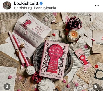 photograph of a copy of The Secret Romantic's Book of Magic, edited by Marie O'Regan and Paul Kane, lying on a wooden platter, on top of scattered pages from books. To the left are pink and white flowers, two pink candles tied with a gold ribbon, and the press release for the book. To the right are a red flower ornamenet, a lit candle in a glass jar, and a Secret Romantic's bookmark