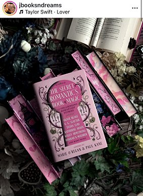 photograph of a copy of The secret Romantic's Book of Magic, edited by Marie O'Regan and Paul Kane, lying on top of a row of hardback books with pink sprayed edges. At the top of the picture is an open book, and the background is various flowers and dark green leaves