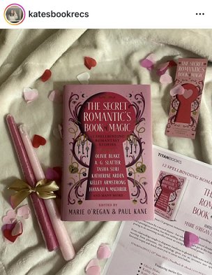 photograph showing a copy of The Secret Romantic's Book of Magic, edited by Marie O'Regan and Paul Kane, lying on a white cloth beside two pink candles tied with a gold ribbon, the press release for the book, a Secret Romantic's bookmark and pink heartshaped confetti