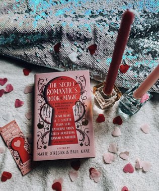 photograph of a copy of The Secret Romantic's Book of Magic, edited by Marie O'Regan and Paul Kane, lying on a white cloth scattered with pink heartshaped confetti. To the left of the book is a Secret Romantic's bookmark, to the right, a dark pink and a light pink candle, both in square glass candleholders. At the top fo the picture is a grey and blue cloth