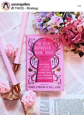 image of a copy of The Secret Romantic's Book of Magic, edited by Marie O'Regan and Paul Kane, lying on a bed of pages from books. To the top right is a bunch of pink and lilac flowers, on the left, two pink candles in brass candlesticks, three pink rose heads