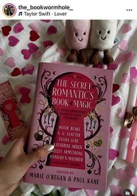 photograph of a woman's hand holding a copy of The Secret Romantic's Book of Magic, edited by Marie O'Regan and Paul Kane, over a white cloth decorated with pink heartshaped confetti. To the left of the book is a Secret Romantic's bookmark, to the right are pink and white candles tied with a gold ribbon. At the top of the picture are a pink and a white felt toy with smiling face
